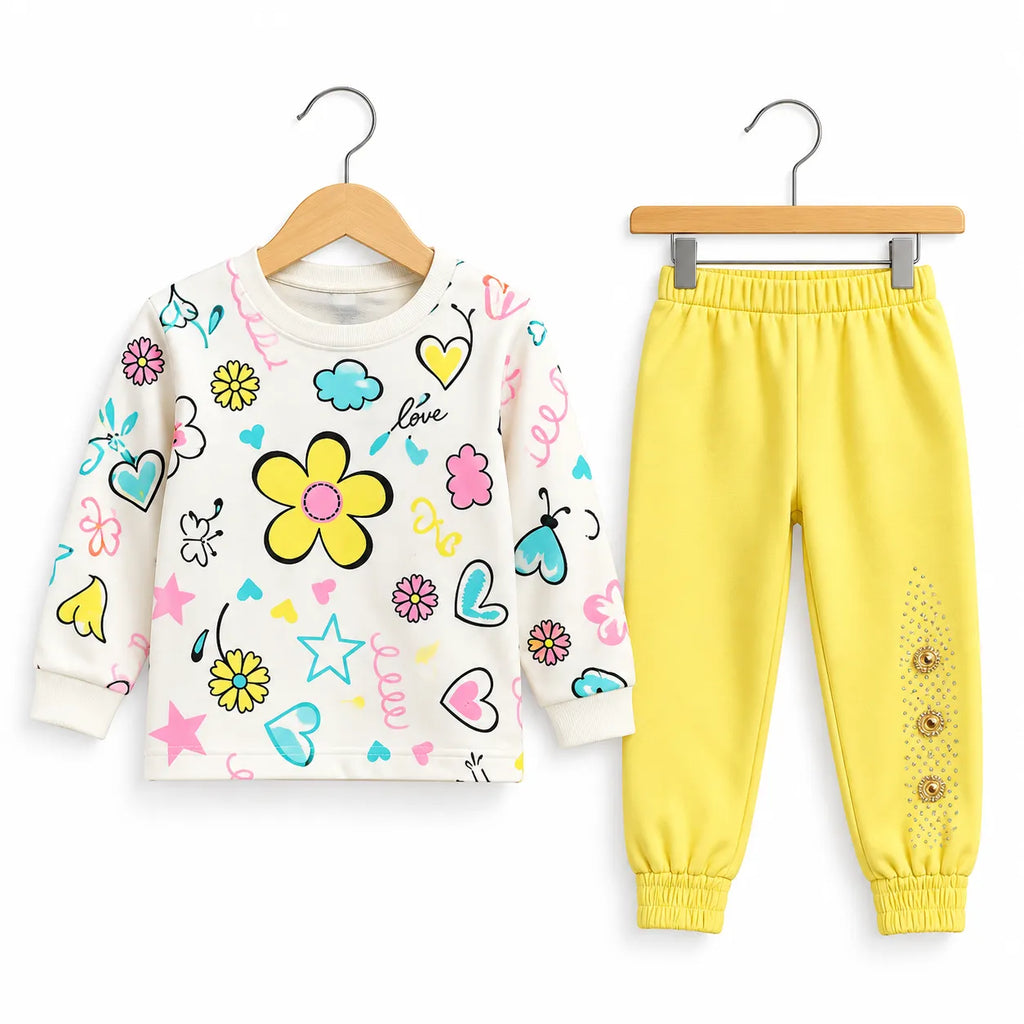 A children's two-piece clothing set on wooden hangers, featuring a cream-colored long-sleeve top with colorful cartoon prints (yellow flower, hearts, stars, butterflies) and matching bright yellow jogger pants with elastic cuffs and decorative buttons.