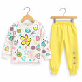 A children's two-piece clothing set on wooden hangers, featuring a cream-colored long-sleeve top with colorful cartoon prints (yellow flower, hearts, stars, butterflies) and matching bright yellow jogger pants with elastic cuffs and decorative buttons.