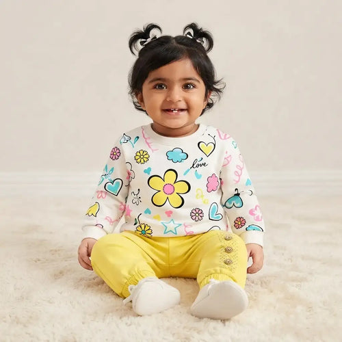 A toddler girl sitting on a white shag rug wearing a white long-sleeve top with colorful cartoon prints and bright yellow pants.