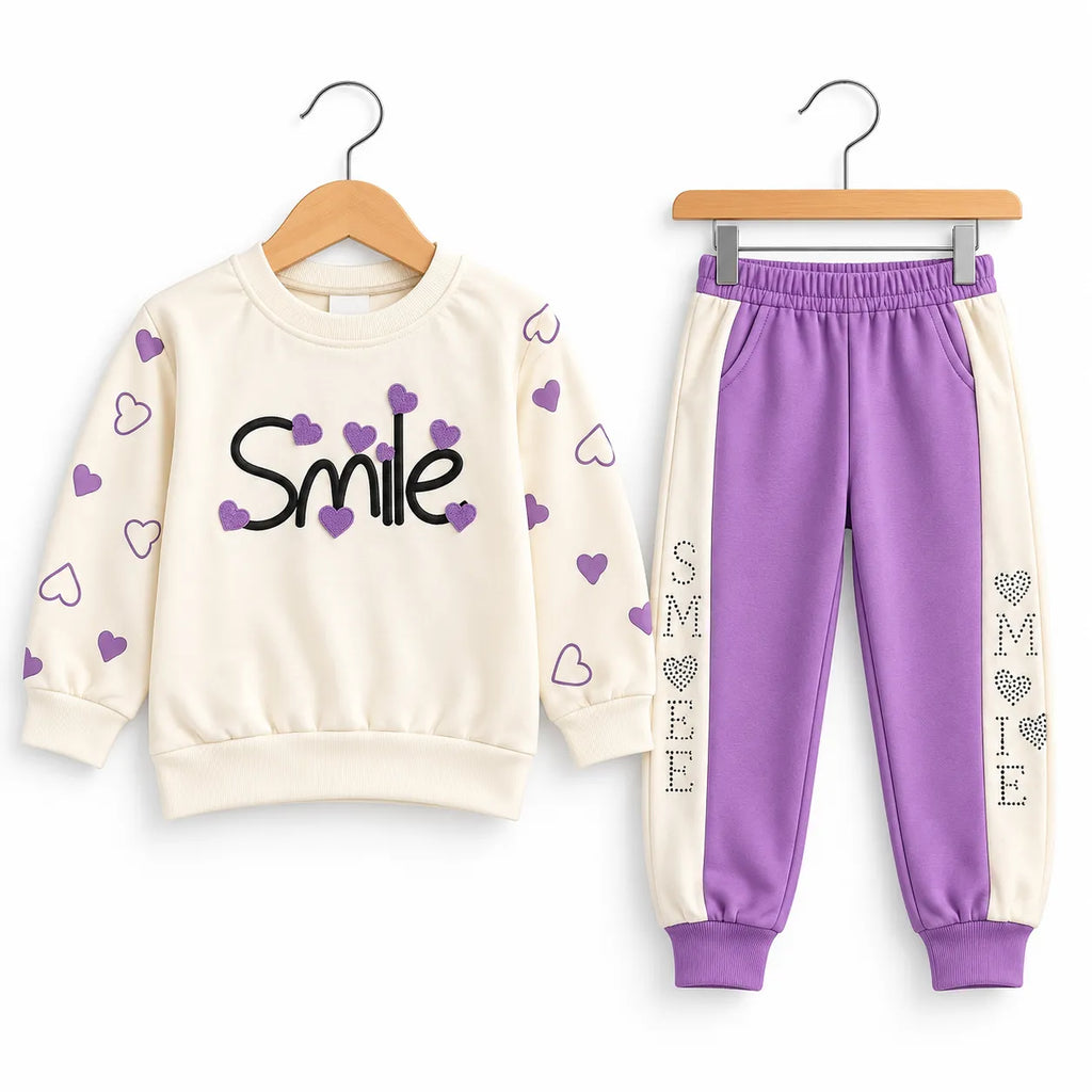 A children's two-piece clothing set featuring a cream sweatshirt with purple hearts and the word 'Smile' in black, paired with matching jogger pants that are purple and cream with 'SMILE' printed vertically on the legs.