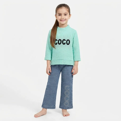 A young girl stands barefoot against a light gray studio background, wearing a mint green textured knit sweater with the word 'COCO' in black across the chest and wide-leg blue jeans with matching 'COCO' lettering down the side.