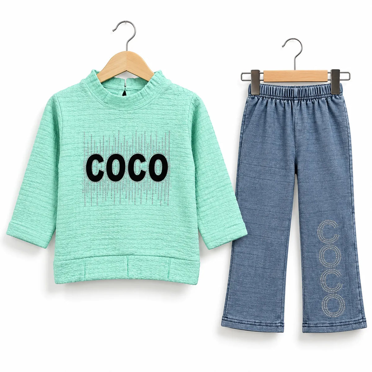 A two-piece children's outfit set on wooden hangers against a white background. The set includes a mint green long-sleeve top with 'COCO' printed in black across the chest, and a pair of blue denim-style pants with 'COCO' printed vertically in circular pattern down one leg.