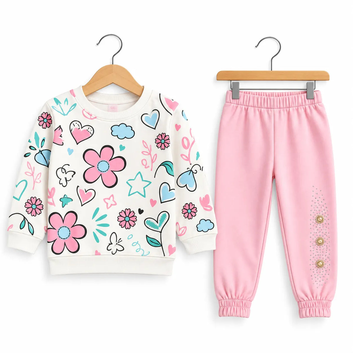 A children's two-piece outfit set featuring a long-sleeve sweatshirt and jogger pants on wooden hangers. The sweatshirt is off-white with a colorful pattern of pink flowers, blue hearts, and teal leaves.