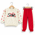 A two-piece children's outfit set on wooden hangers, featuring a cream sweatshirt with red hearts and the word 'Smile' in black, paired with red jogger pants with cream side panels.