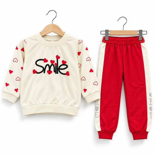 A two-piece children's outfit set on wooden hangers, featuring a cream sweatshirt with red hearts and the word 'Smile' in black, paired with red jogger pants with cream side panels.