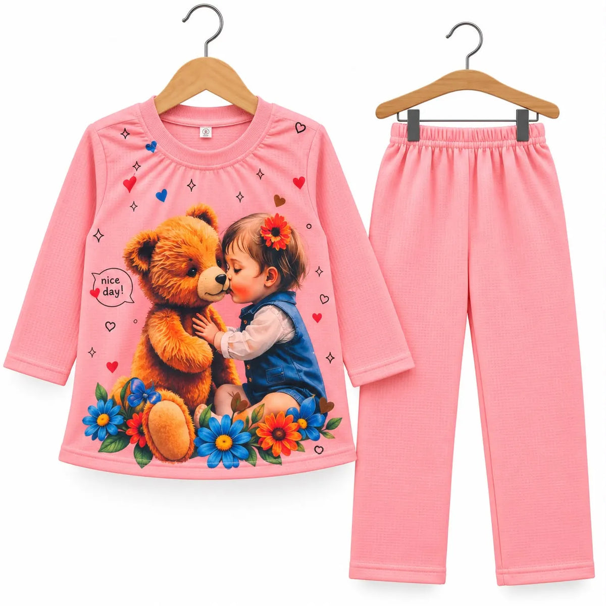 A two-piece children's pajama set consisting of a long-sleeve pink top and matching pants, displayed on wooden hangers against a white background. The top features a colorful graphic print of a young girl kissing a brown teddy bear, surrounded by blue and orange flowers, with small heart and star patterns scattered across the fabric.