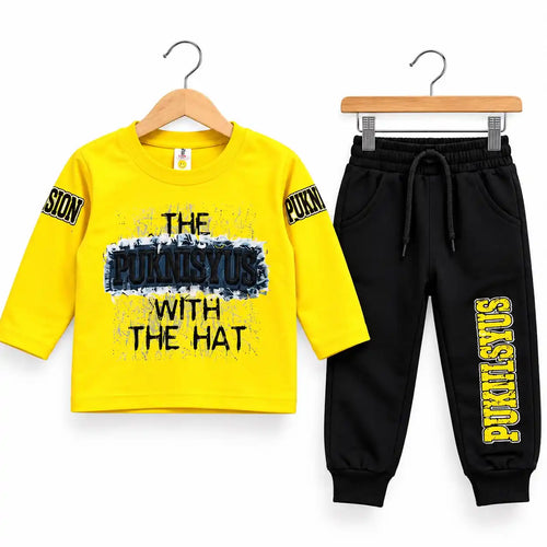 A children's two-piece outfit set on wooden hangers against a white background. The set includes a bright yellow long-sleeve t-shirt with a black and blue graphic print reading 'THE PUKNSYUS WITH THE HAT' and black printed sleeve patches, paired with black jogger pants featuring a drawstring waist and 'PUKNYSYUS' printed in yellow down one leg.