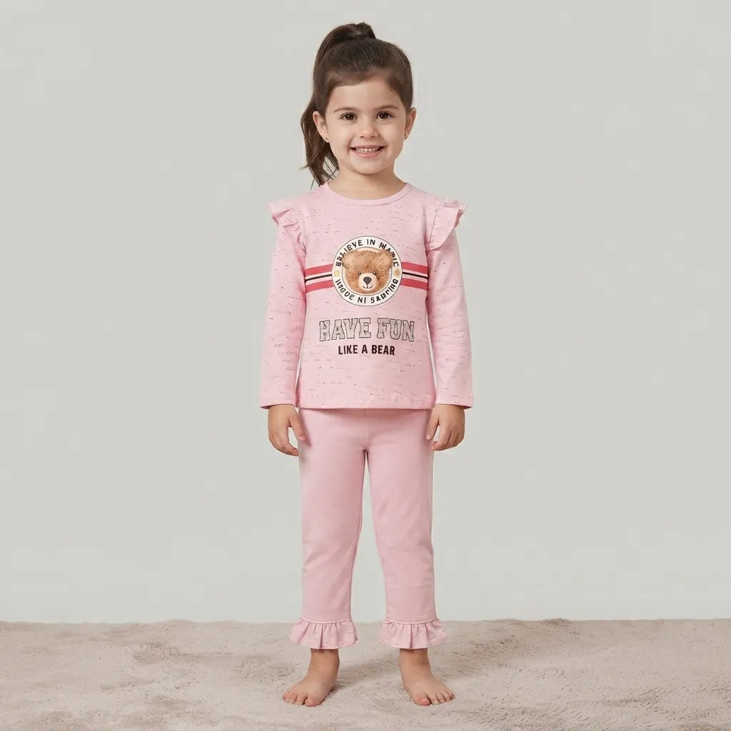 A young girl stands barefoot against a light gray studio background wearing a pink two-piece pajama set with a teddy bear graphic on the top and ruffled cuffs on the pants.
