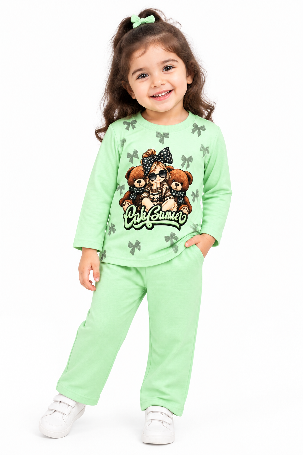 A young girl with brown hair in a ponytail wearing a mint green two-piece pajama set with a teddy bear graphic and white sneakers, standing against a plain white background.